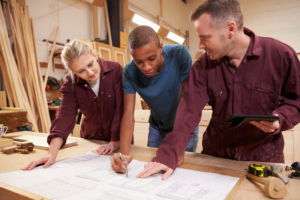 Carpenter With Apprentices Looking At Plans In Workshop